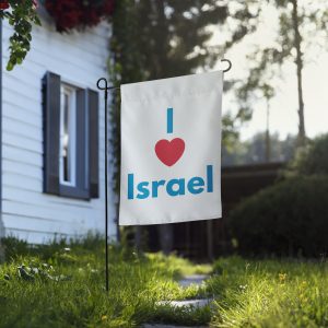 A white garden flag displayed outside a house, featuring the text “I Love Israel” in blue and red, standing on a lawn with greenery and a sunlit background.