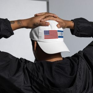 Back view of a person wearing a white cap with embroidered United States and Israeli flags, holding the cap with both hands.