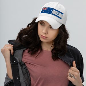 A woman wearing a white cap featuring embroidered Australia and Israel flags, paired with a casual outfit, looking to the side against a neutral background